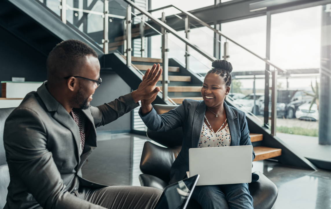 Two professionals in business attire high-fiving while seated in a modern office space.