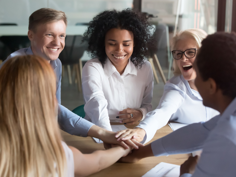 Group of diverse professionals engaged in a collaborative meeting, smiling and joining hands on a table, symbolizing teamwork and unity.