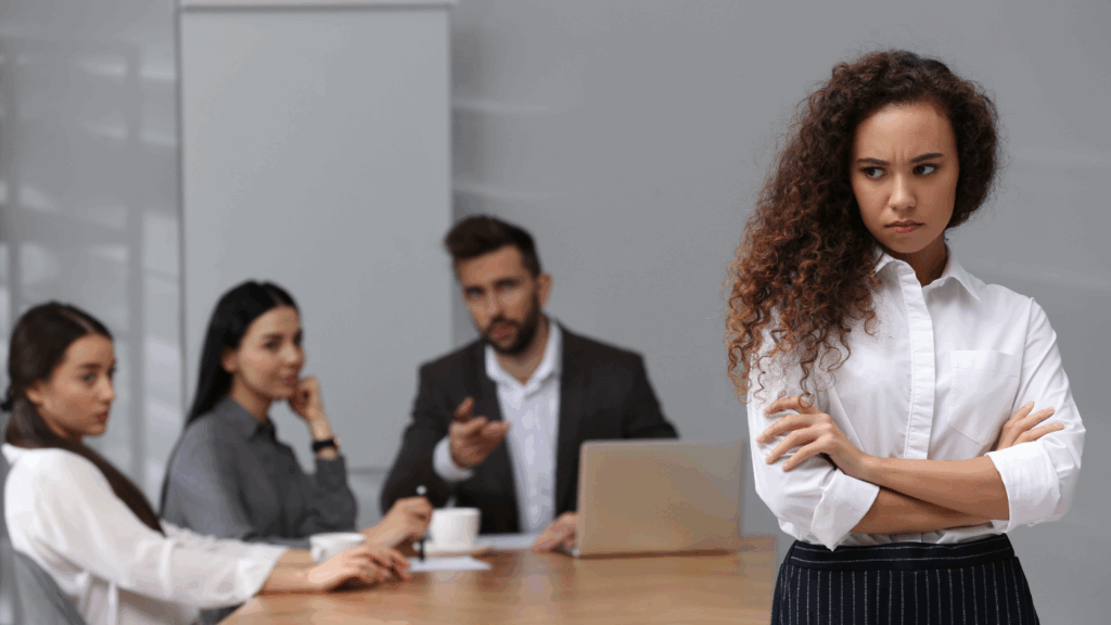 A woman with curly hair stands with her arms crossed in a meeting room while others are seated at a table.