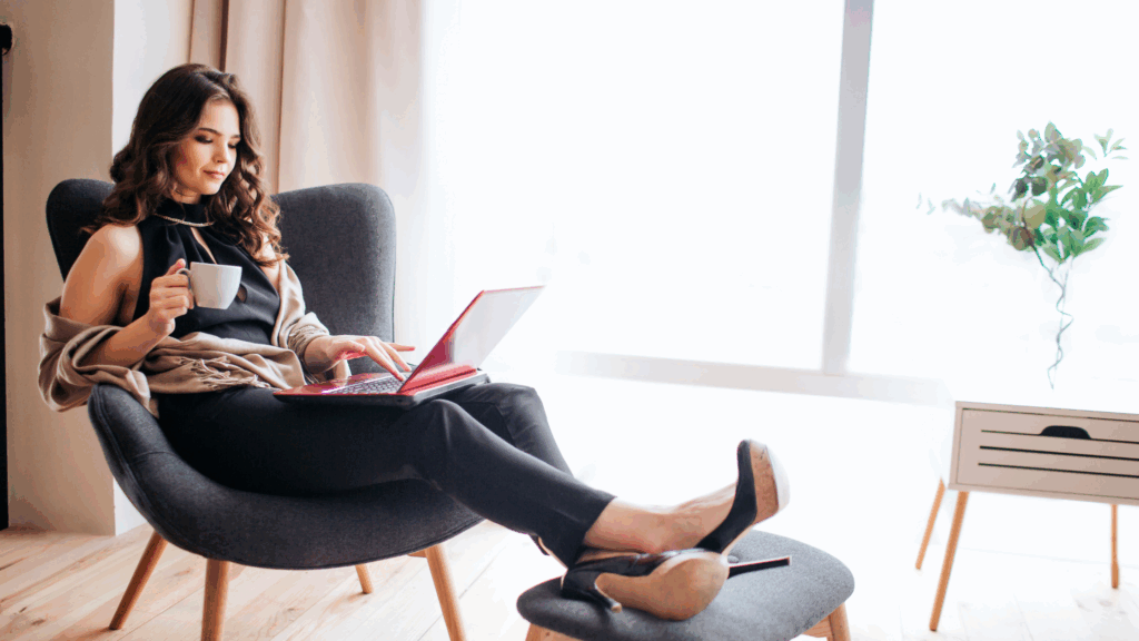 Woman sitting in a gray chair using a laptop while holding a cup in a well-lit room.