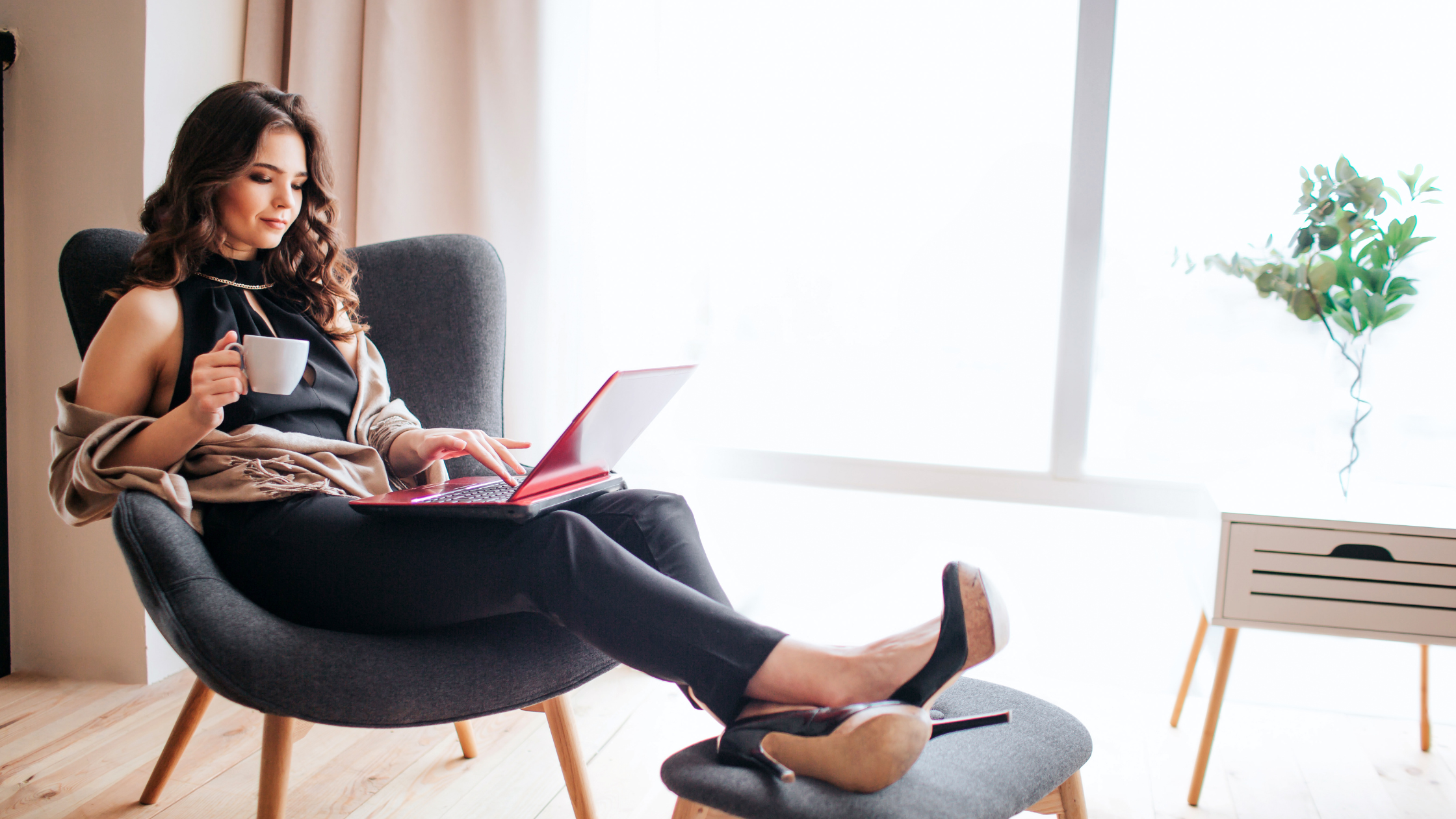 Woman sitting in a gray chair using a laptop while holding a cup in a well-lit room.