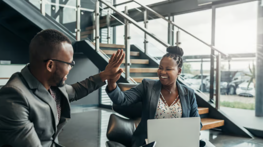 Two business professionals smiling and giving a high five while seated in an office environment.