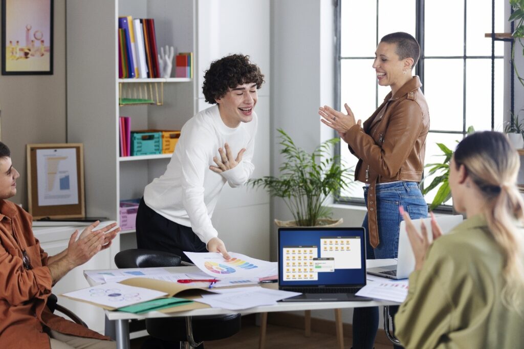 The Atrium, LLC team in a bright open-plan office, one person standing and clapping with others seated, papers and laptops on the table