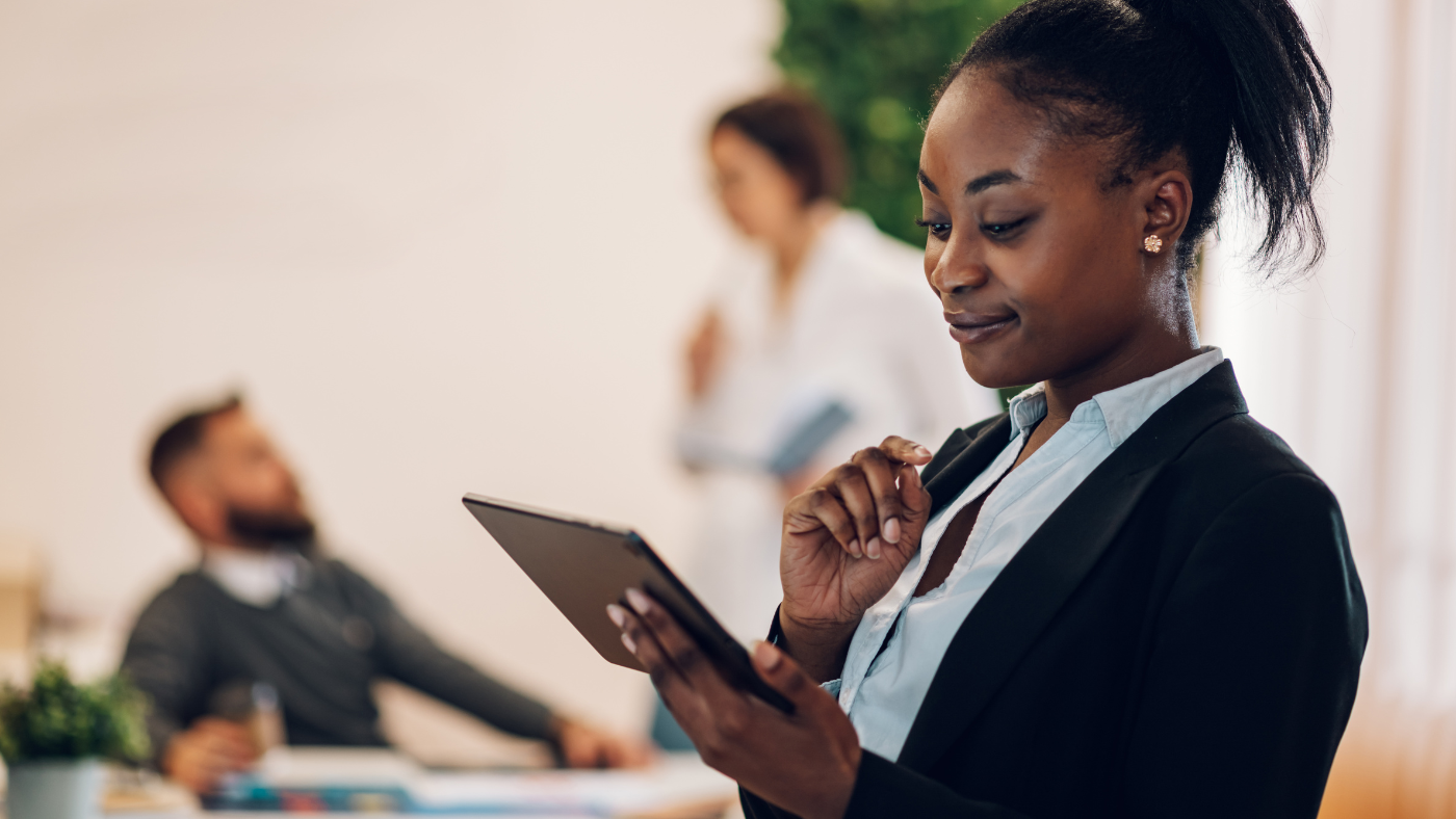 Professional Black woman in a black blazer and light blue blouse holding a tablet in an office setting, with colleagues blurred in the background.