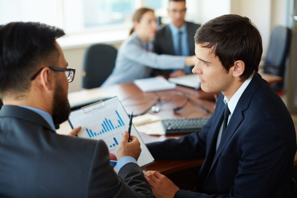Two men in dark suits discuss a chart on a clipboard while colleagues in business attire work at a conference table, reflecting an executive coaching context in financial services.