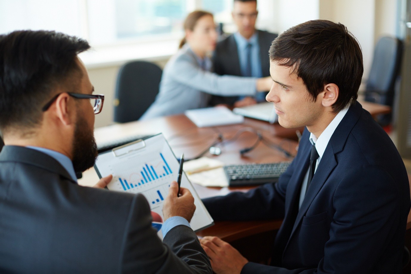 Two men in dark suits discuss a chart on a clipboard while colleagues in business attire work at a conference table, reflecting an executive coaching context in financial services.