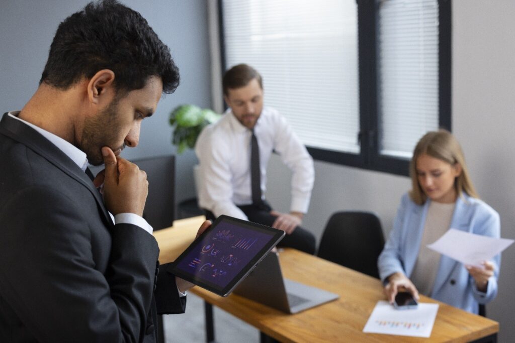 The Atrium, LLC executives in a modern office review data on a tablet with charts; a man in a dark suit stands near a wooden table while two colleagues review documents and a laptop sits on the table