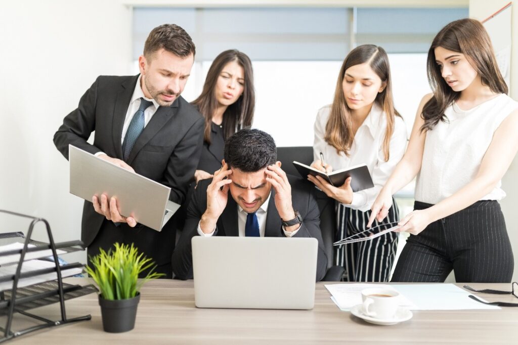 Stressed executive at a desk with laptop surrounded by HR team in a busy office, discussing hiring strategy; The Atrium, LLC