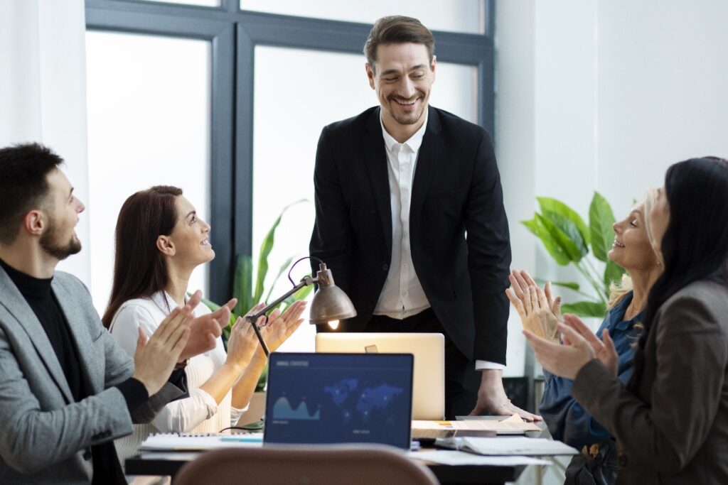 A group of professionals in business attire applaud as a smiling man in a black suit stands at a conference table in a bright office, suggesting a workplace meeting or presentation.