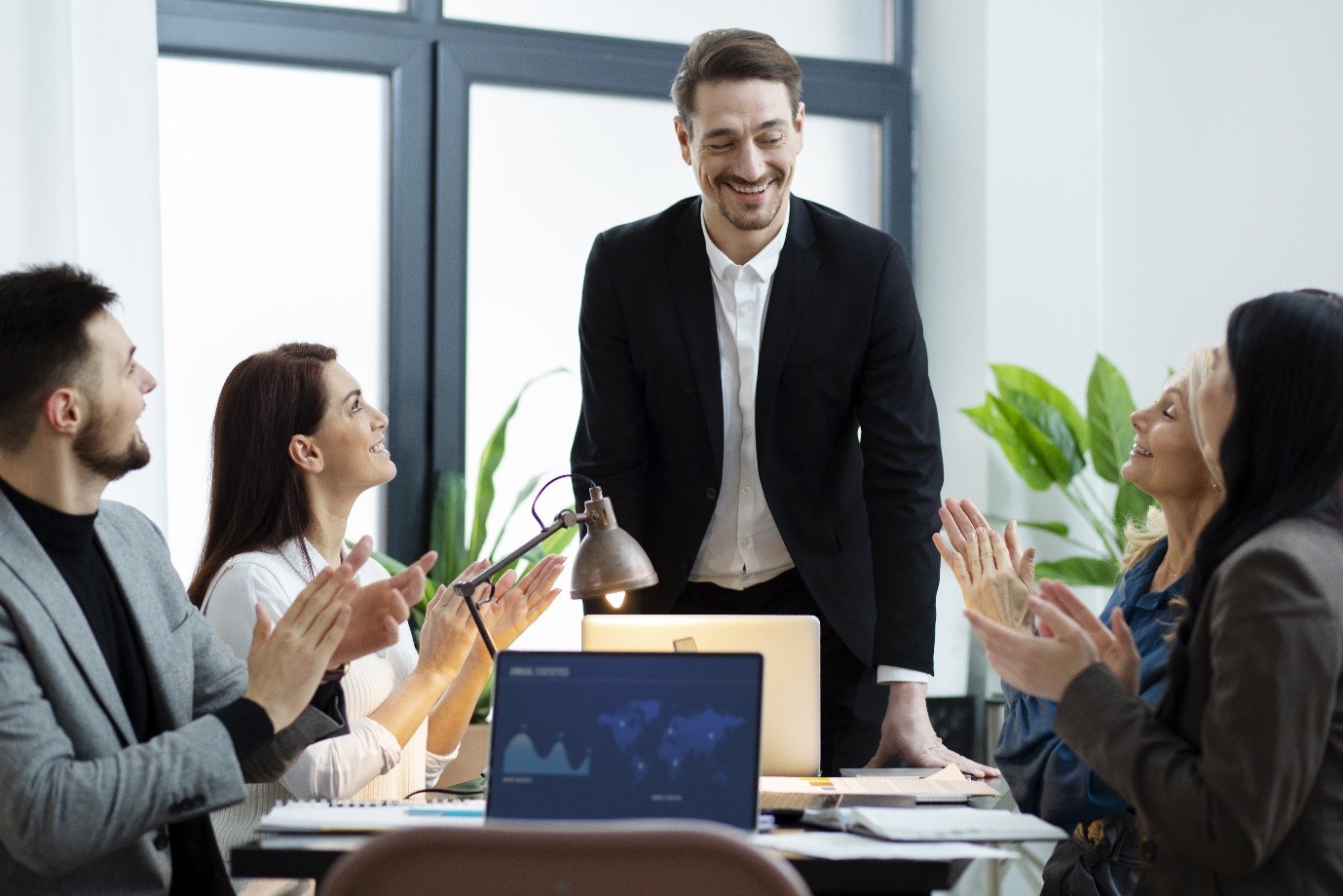 A group of professionals in business attire applaud as a smiling man in a black suit stands at a conference table in a bright office, suggesting a workplace meeting or presentation.