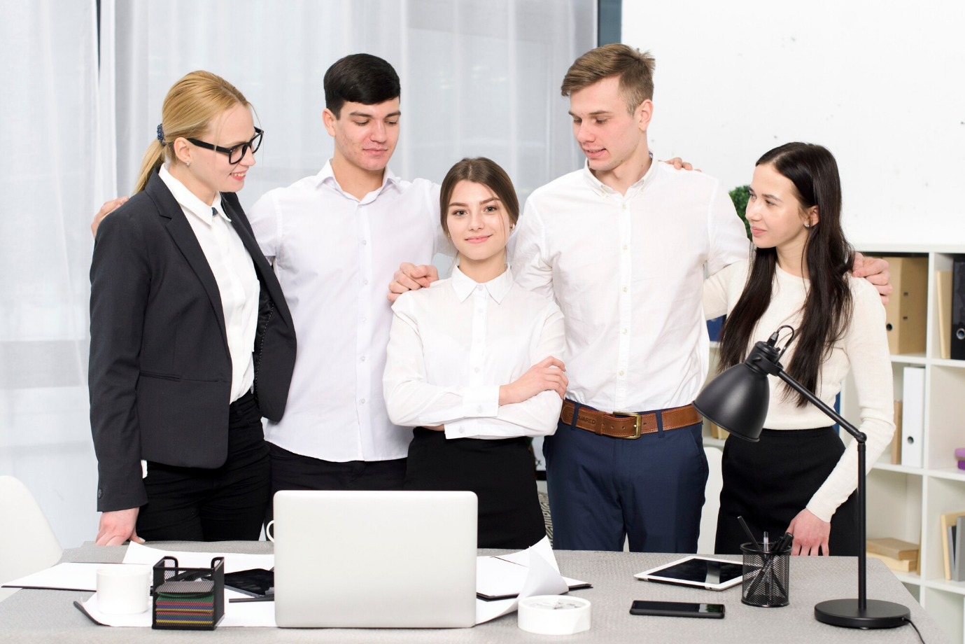 Five young professionals in business attire standing together around a desk with a laptop and documents.